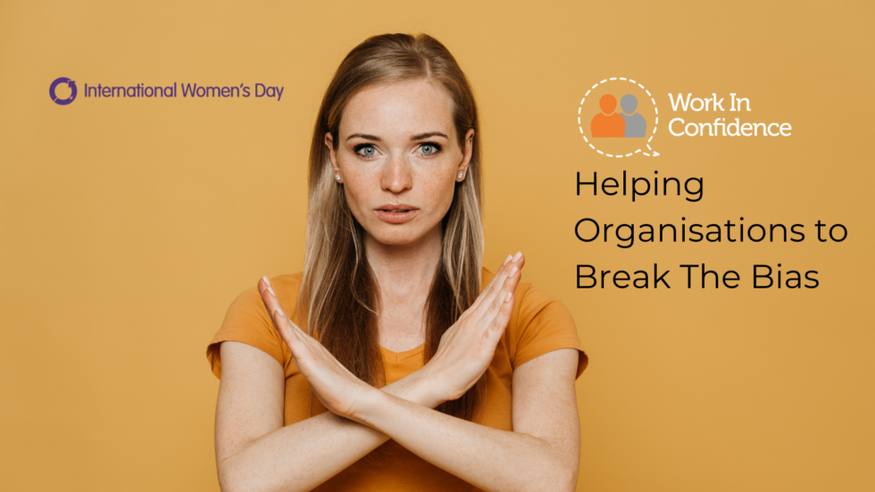 A white woman wearing a mustard top with hands crossed a shoulder height with the text "helping organisations to Break the Bias" and the International Women's Day logo