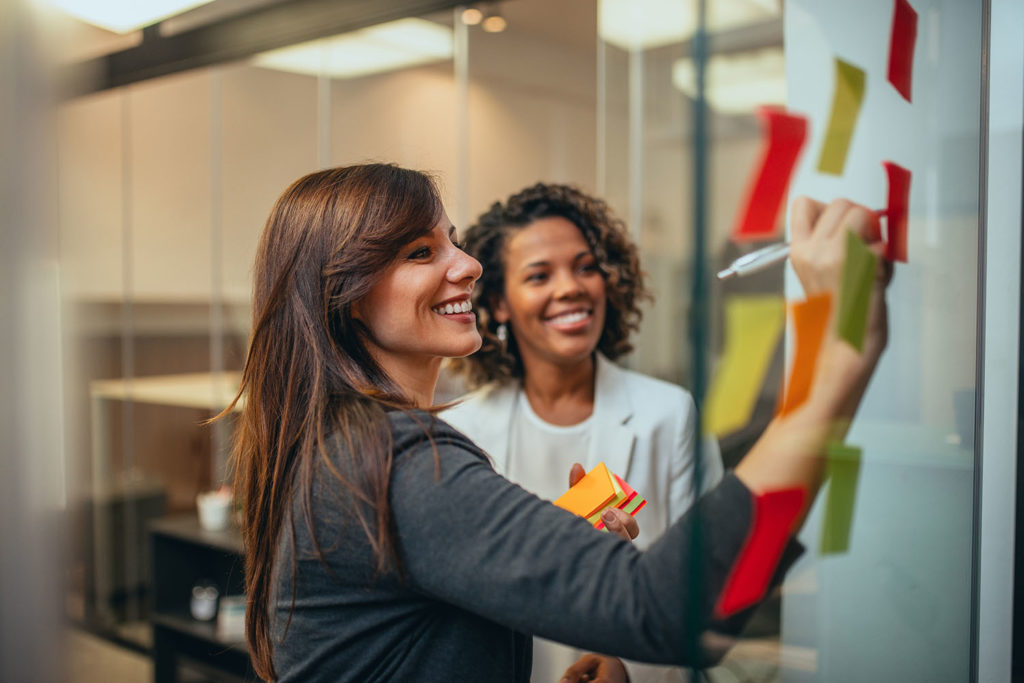 Smiling businesswomen brainstorming with adhesive notes on a glass wall in the office.